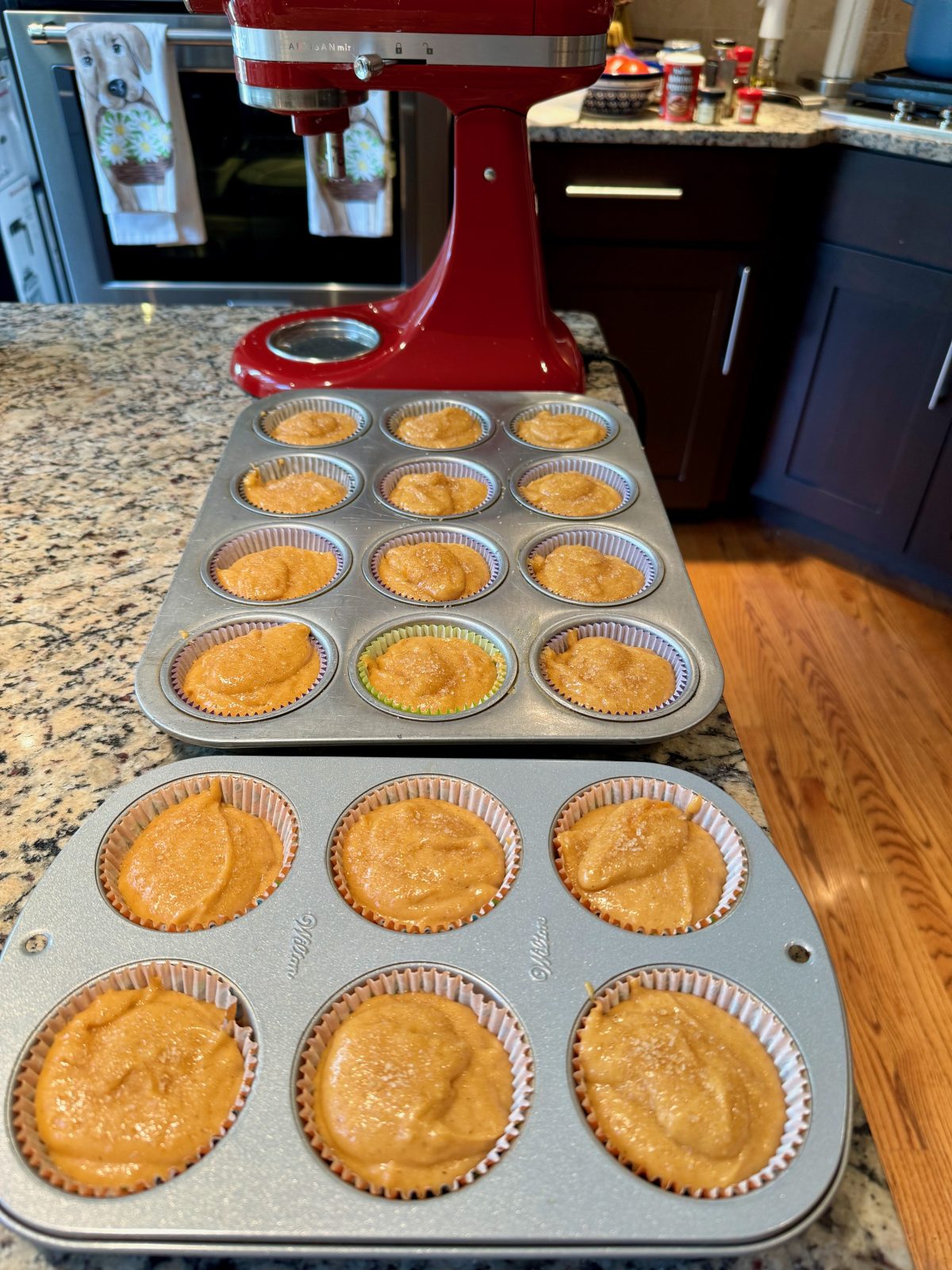 18 unbaked pumpkin muffins in 12- cup and 6-cup muffin pans on a kitchen counter with a red KitchenAid stand mixer in the background.