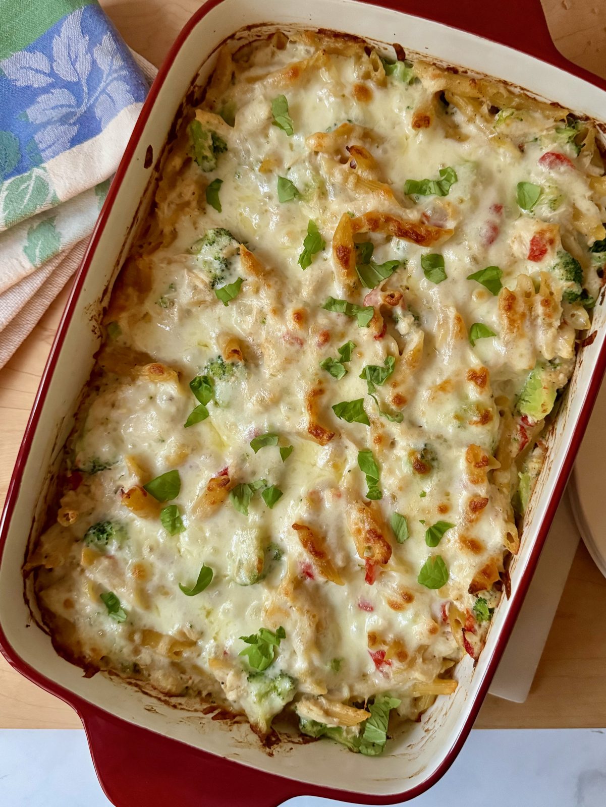 Red 13x9 stoneware casserole with broccoli, chicken, red pepper, penne pasta casserole topped with melted and browned mozzarella cheese and sprinkled with fresh basil.