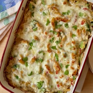 Red 13x9 stoneware casserole with broccoli, chicken, red pepper, penne pasta casserole topped with melted and browned mozzarella cheese and sprinkled with fresh basil.