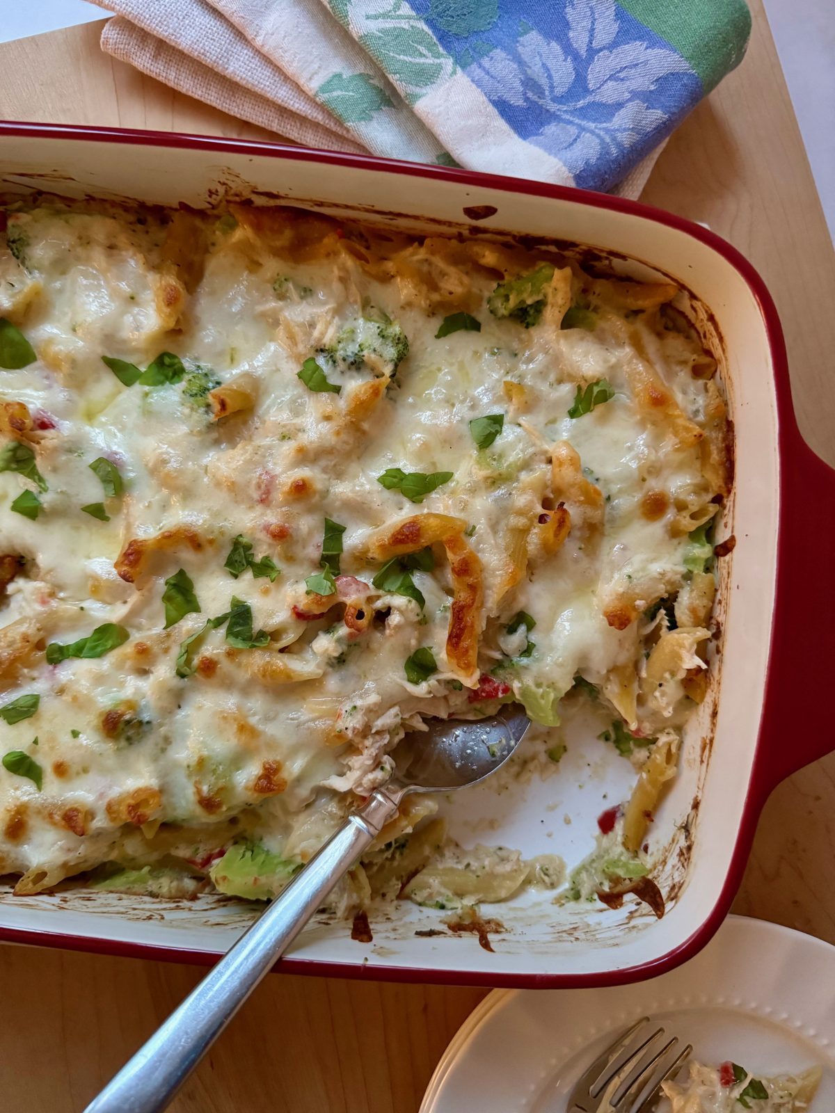 Red 13x9 stoneware casserole with broccoli, chicken, red pepper, penne pasta casserole topped with melted and browned mozzarella cheese and sprinkled with fresh basil.  One portion is spooned out and you can see the edge of a cloth napkin and plate.