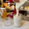 Pouring thick tan opaque colored whipped honey from a glass KitchenAid Stand Mixer Bowl into a mason jar. Mixer and honey jar in background