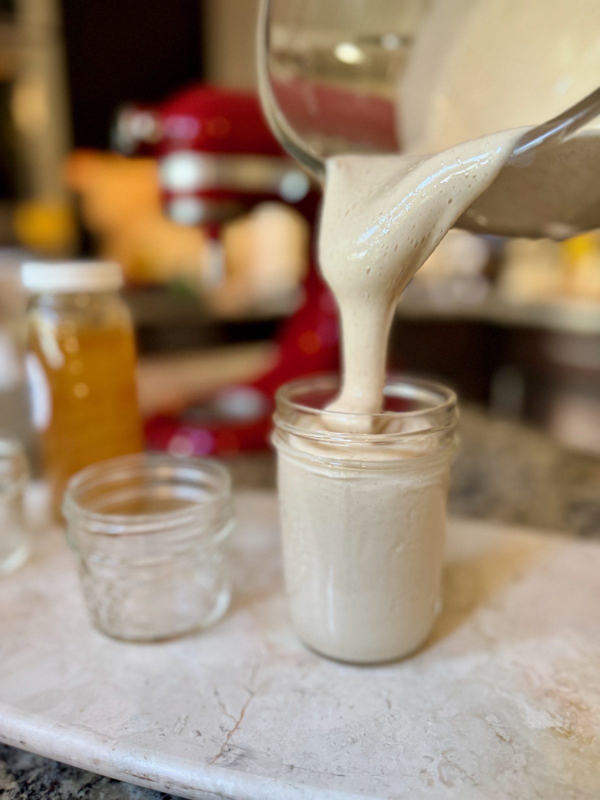 Pouring thick tan opaque colored whipped honey from a glass KitchenAid Stand Mixer Bowl into a mason jar. Mixer and honey jar in background