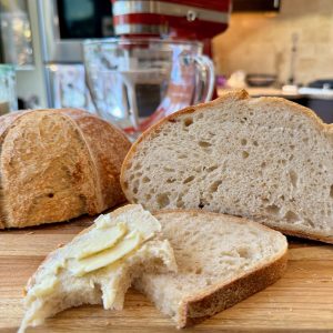 Homemade Sourdough bread sliced exposing airy interior of loaf. One slice cut and slathered with butter. A KitchenAid Artisan Mini 3.5 qt Stand Mixer with glass bowl in the background.