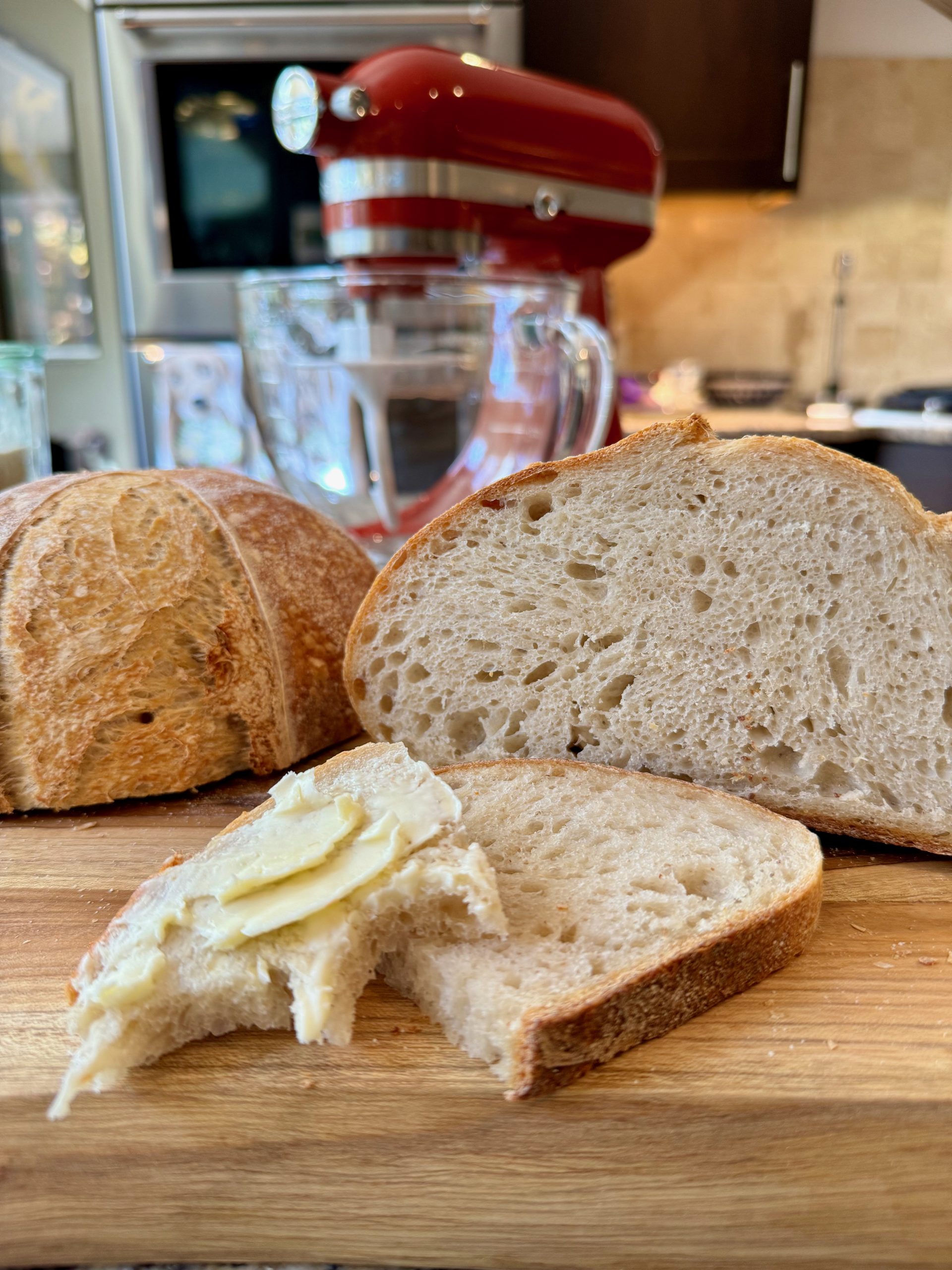 Homemade Sourdough bread sliced exposing airy interior of loaf. One slice cut and slathered with butter. A KitchenAid Artisan Mini 3.5 qt Stand Mixer with glass bowl in the background.