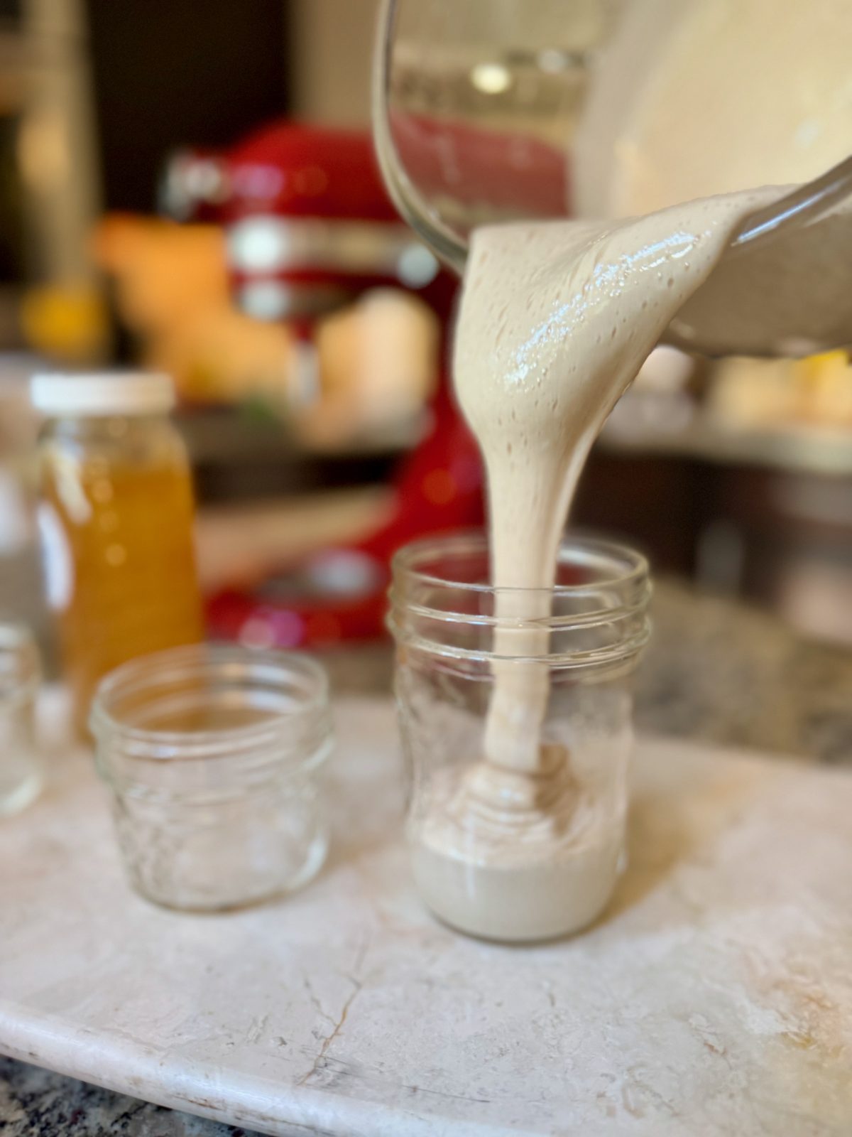 Pouring thick tan opaque colored whipped honey from a glass KitchenAid Stand Mixer Bowl into a mason jar.  Mixer and honey jar in background