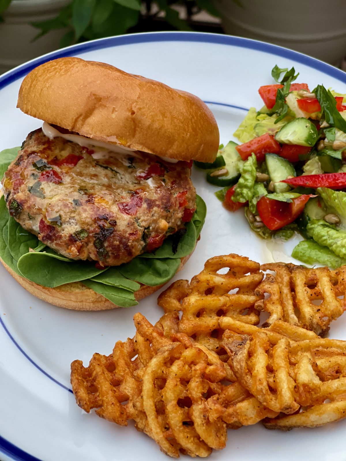 Cooked golden brown turkey burger looks juicy with flecks of red pepper, feta and spinach on a brioche bun with baby spinach. Plate also has sweet potato waffle fries and a simple chopped salad with tomato and cucumber