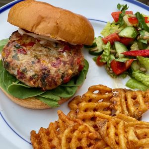 Cooked golden brown turkey burger looks juicy with flecks of red pepper, feta and spinach on a brioche bun with baby spinach. Plate also has sweet potato waffle fries and a simple chopped salad with tomato and cucumber