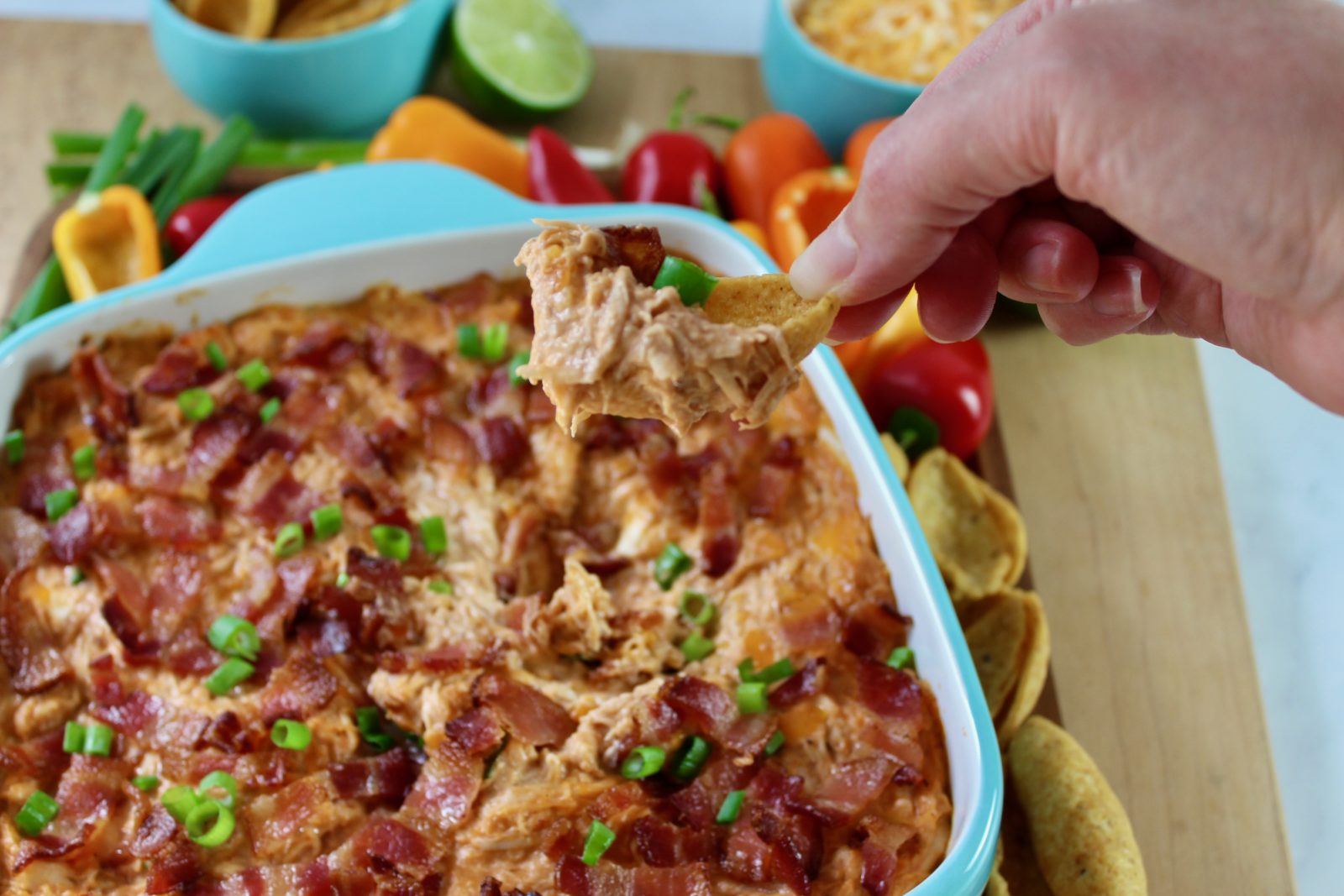 Square turquoise blue baking dish with freshly baked BBQ Chicken Dip.  Dip is topped with loads of bacon and a sprinkle of green onion.  Baking dish is surrounded by colorful peppers, Fritos Scoops and some shredded cheddar in a matching blue bowl.  There is also a half of juicy lime.  A hand is scooping some dip with a Frito Scoop chip.