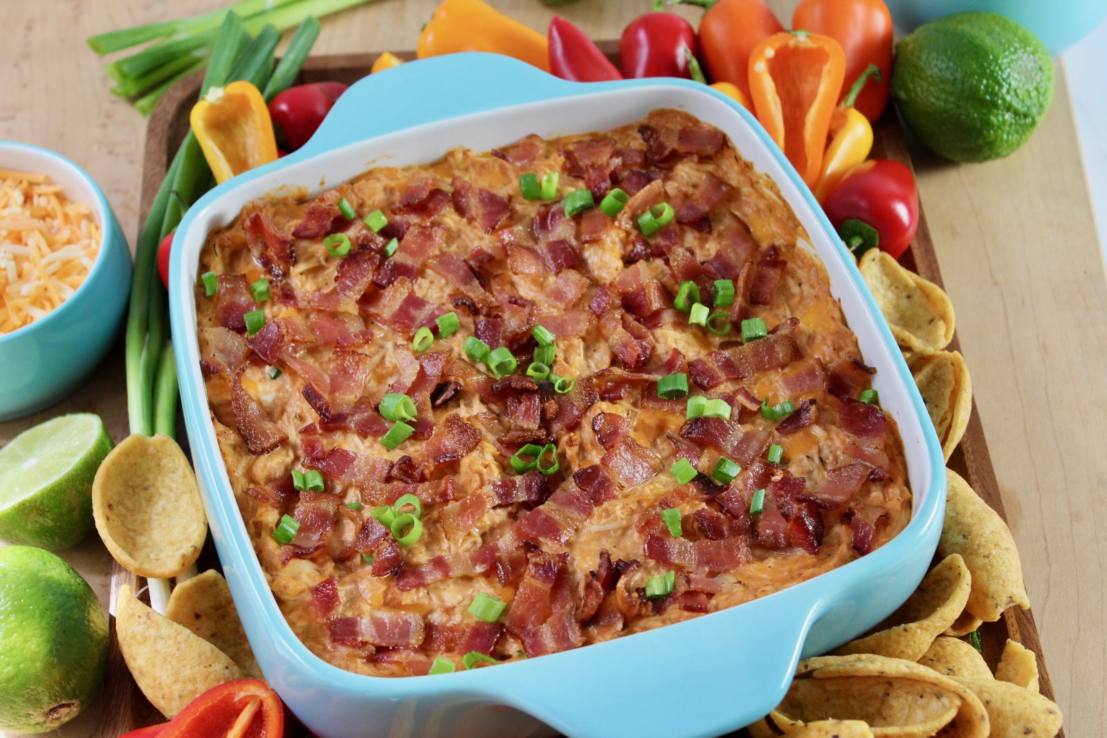 A landscape oriented recipe photo. Square turquoise blue baking dish with freshly baked BBQ Chicken Dip.  Dip is topped with loads of bacon and a sprinkle of green onion.  Baking dish is surrounded by colorful peppers, Fritos Scoops and some shredded cheddar in a matching blue bowl.  There is also a half of juicy lime.