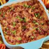 Square turquoise blue baking dish with freshly baked BBQ Chicken Dip. Dip is topped with loads of bacon and a sprinkle of green onion. Baking dish is surrounded by colorful peppers, Fritos Scoops and some shredded cheddar in a matching blue bowl. There is also a half of juicy lime.