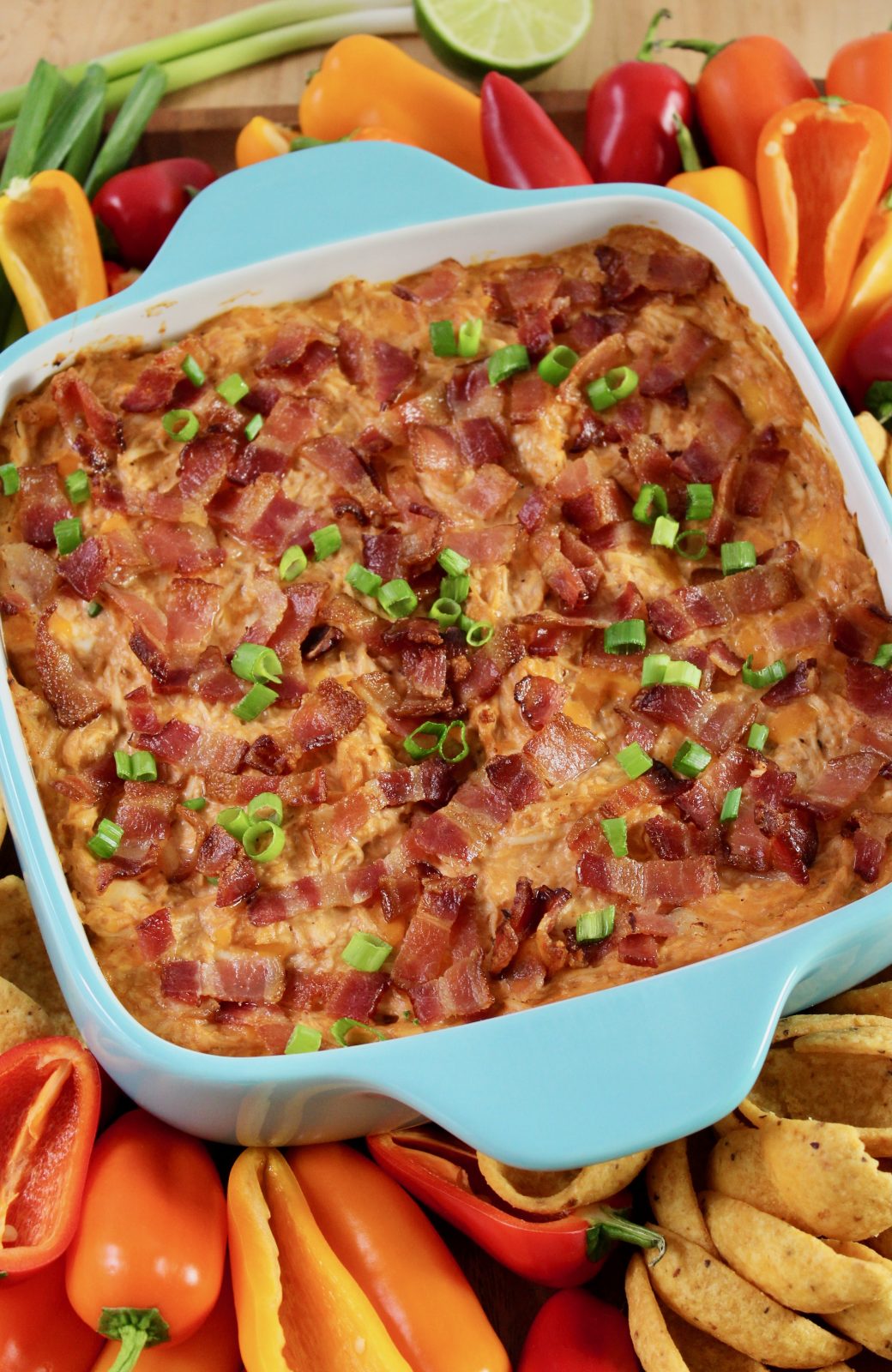 Square turquoise blue baking dish with freshly baked BBQ Chicken Dip. Dip is topped with loads of bacon and a sprinkle of green onion. Baking dish is surrounded by colorful peppers, Fritos Scoops and some shredded cheddar in a matching blue bowl. There is also a half of juicy lime.