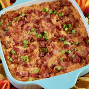 Square turquoise blue baking dish with freshly baked BBQ Chicken Dip. Dip is topped with loads of bacon and a sprinkle of green onion. Baking dish is surrounded by colorful peppers, Fritos Scoops and some shredded cheddar in a matching blue bowl. There is also a half of juicy lime.