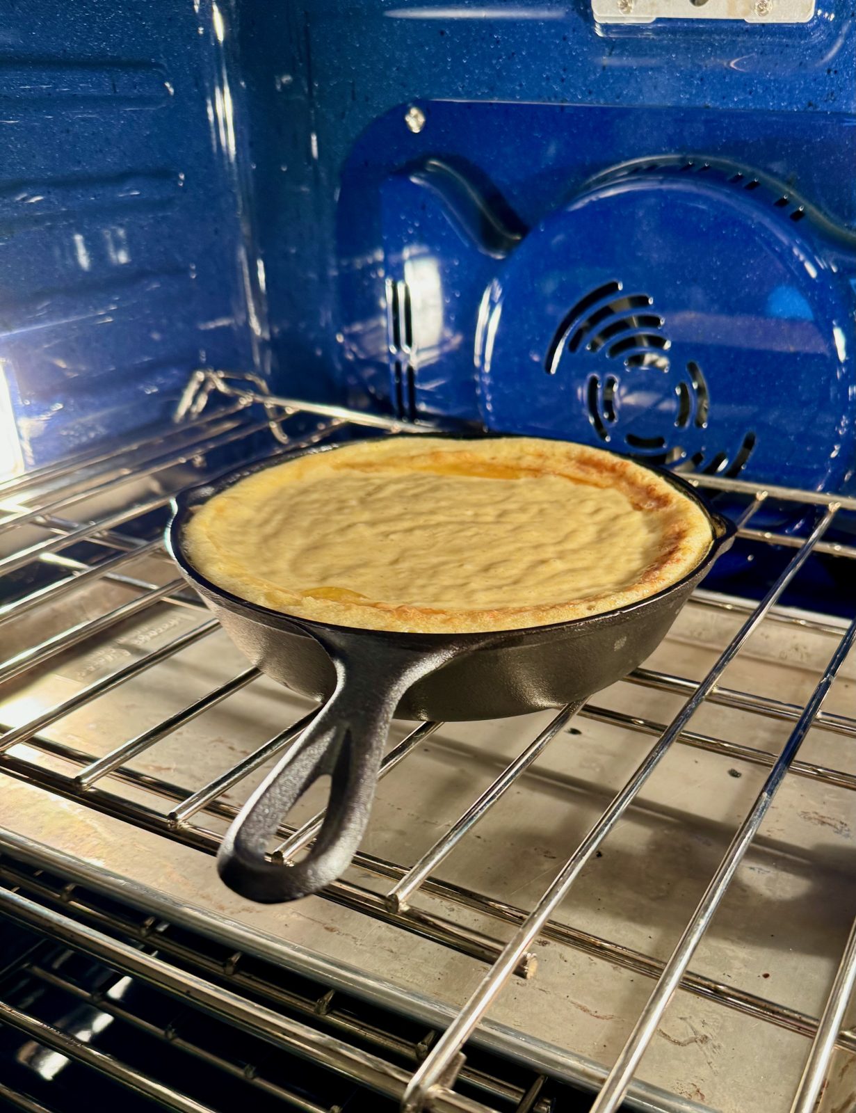 recipe photo for sourdough cornbread showing cornbread batter baking in oven with blue interior.  the edges of the cornbread are puffed up inside the 8-inch cast iron skillet